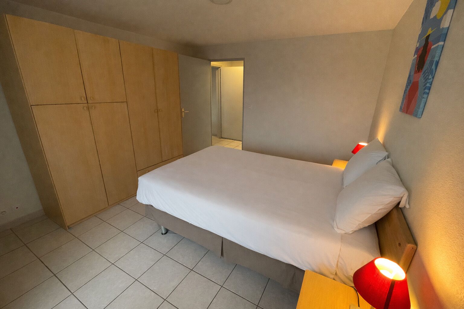Un dormitori amb un llit, un armari, una tauleta de nit amb una làmpada, i una porta que dona a una altra habitació.