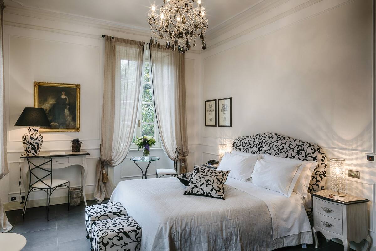 A bedroom with a large bed, a desk, a chair, a nightstand, and a chandelier.