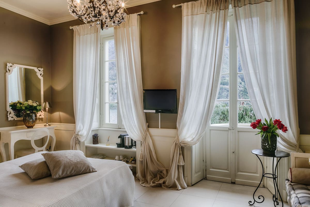 A bedroom with a bed, a TV, a window with curtains, a small table with a vase, and a chair.