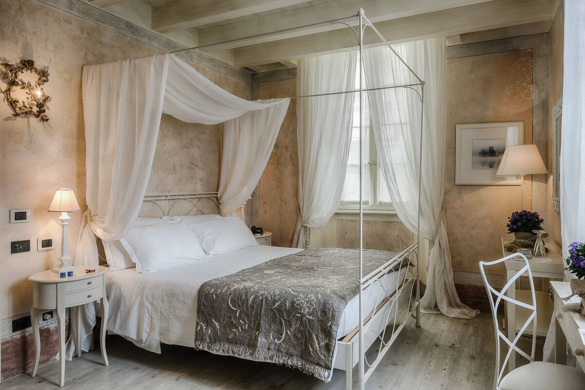 A bedroom with a four-poster bed, a bedside table with a lamp, a chair, and a small table with a vase.
