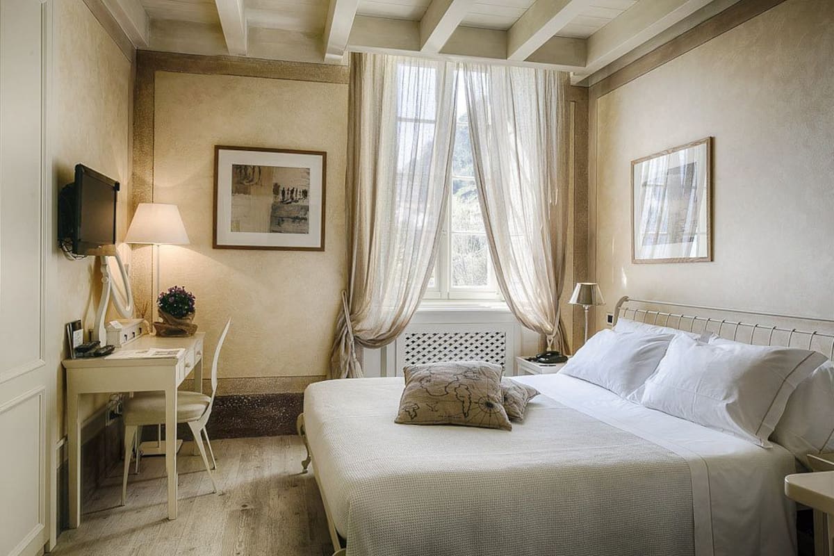 A bedroom with a bed, a desk, a chair, a TV, and two windows with sheer curtains.