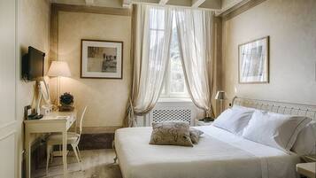 Un dormitorio con una cama, un escritorio, una silla, un televisor y dos ventanas con cortinas translúcidas.