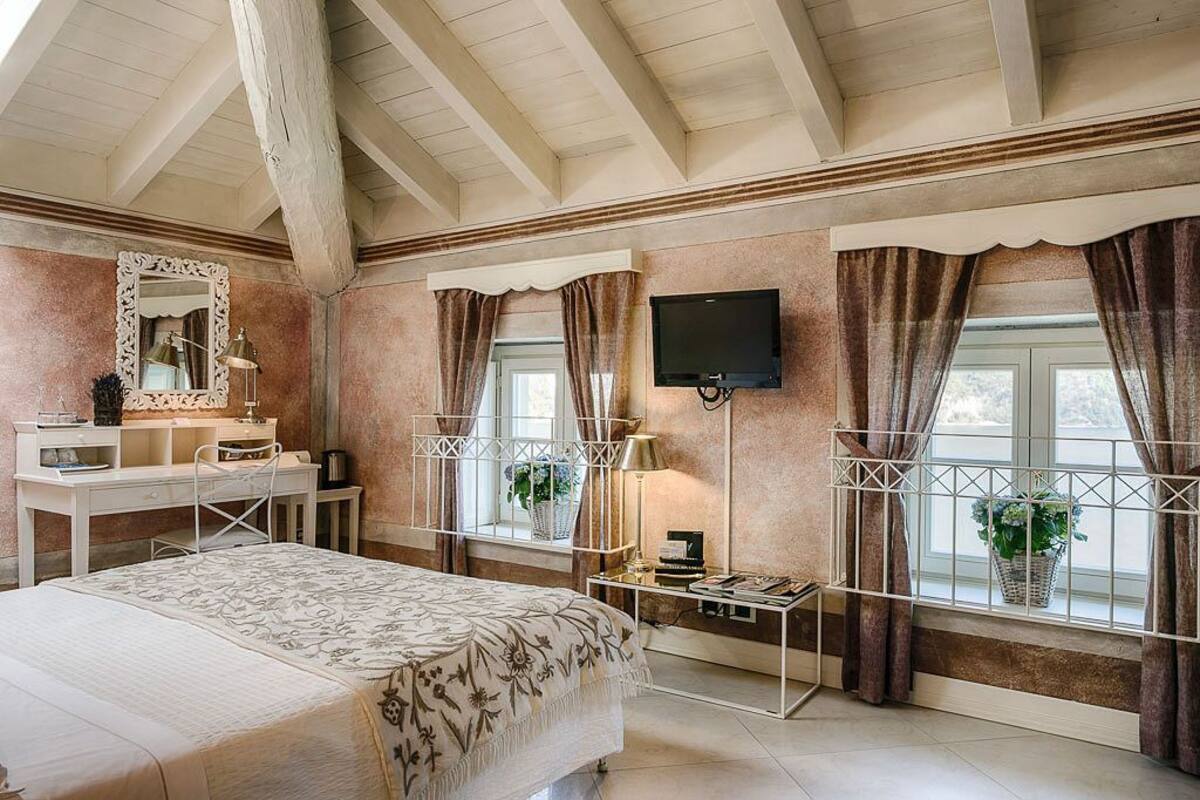 A bedroom with a bed, a desk, a mirror, a TV, and windows with curtains.