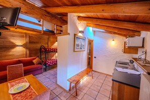 A cozy room with a wooden ceiling, a sofa, a bench, a table with a plate, and a wall-mounted TV. - Residence Trompe-L'Oeil (Ayas)