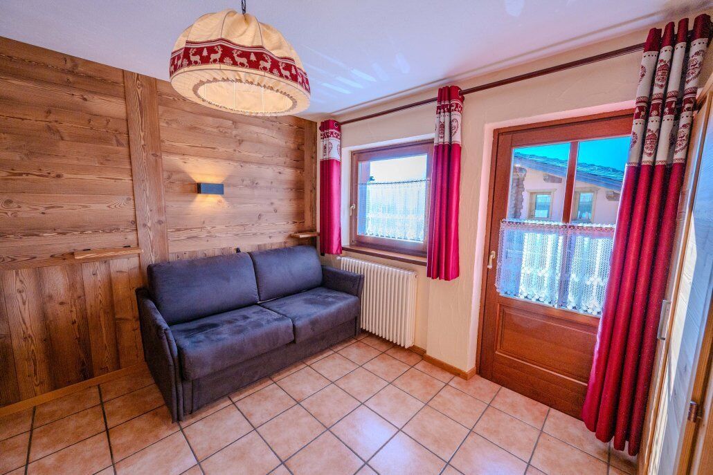 A room with a wooden wall, a black sofa, a window with red curtains, and a door with patterned curtains.