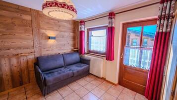 A room with a wooden wall, a black sofa, a window with red curtains, and a door with patterned curtains.
