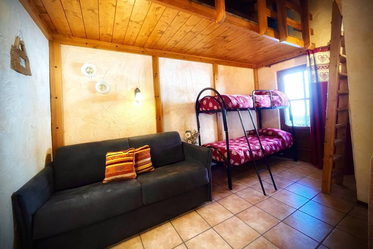 Una habitación acogedora con literas, un sofá con cojines estampados, vigas de madera en el techo y una ventana con cortinas.