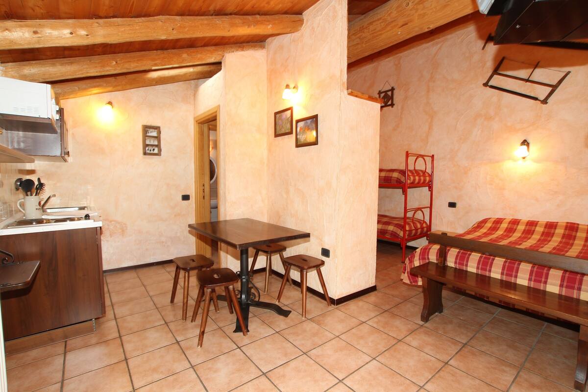 Una habitación acogedora con una mesa de madera, dos taburetes, una cama litera con manta a cuadros y una cocina con un fregadero y armarios.