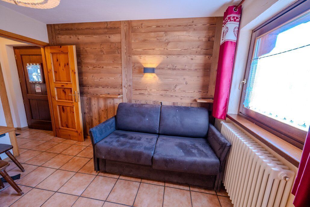 A room with a wooden wall, a window with a red curtain, a radiator, and a dark-colored sofa.