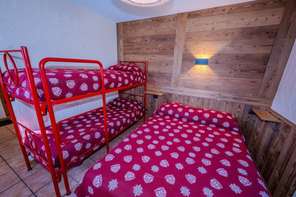 Una cama litera con estructura metálica roja y ropa de cama estampada, un panel de pared de madera y una lámpara fijada a la pared.
