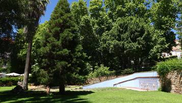 Un jardín bien cuidado con diversos árboles, una zona pavimentada y un muro decorativo, bajo un cielo azul despejado.
