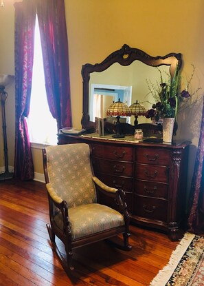 Quarto com uma cadeira de madeira, cômoda com gavetas, espelho, abajur e um vaso com flores.
