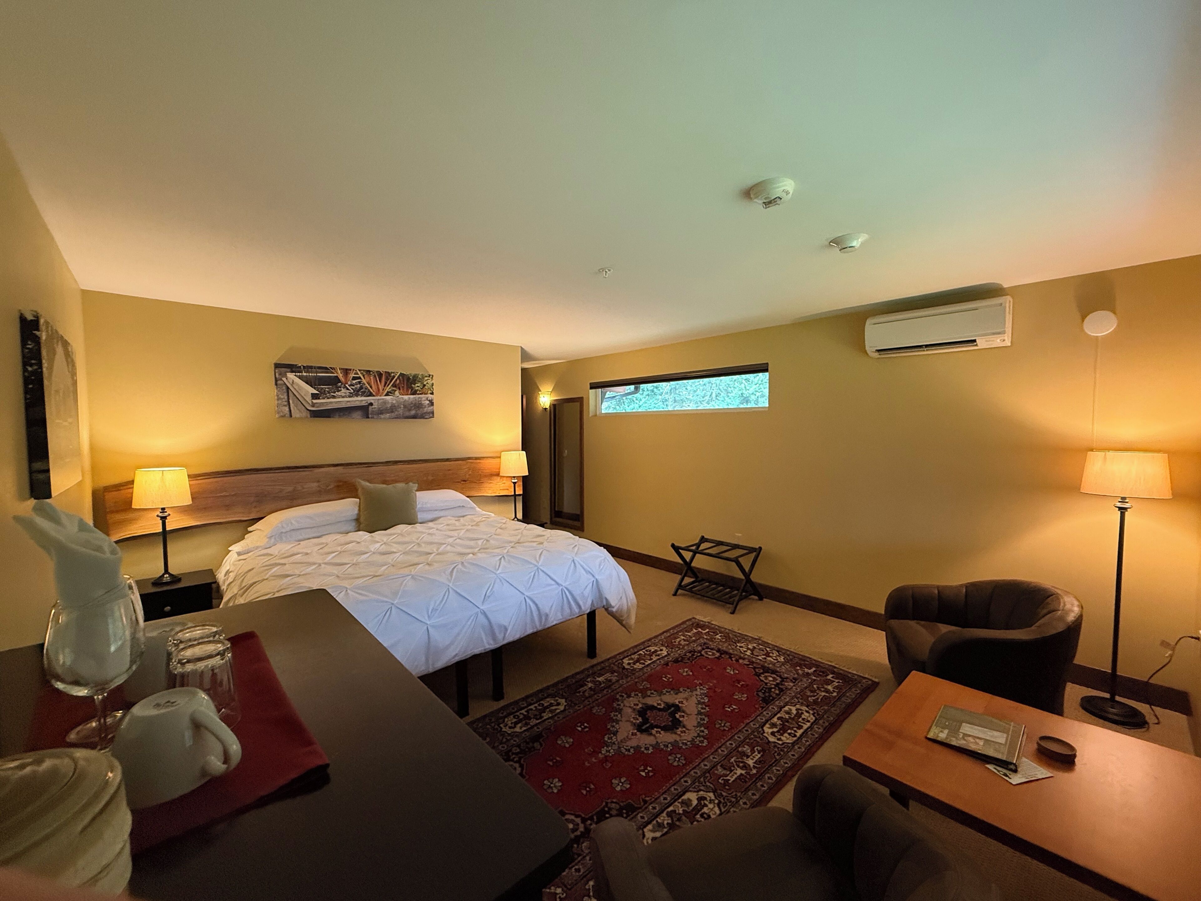 A hotel room with a bed, two bedside lamps, a small table, a chair, a red rug, a window with a view, and an air conditioning unit.
