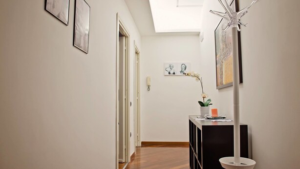 A hallway with a wooden floor, white walls, and a ceiling light. There are framed pictures on the walls, a white vase with an orchid, and a white coat stand. - B&B Sul Corso (Salerno)