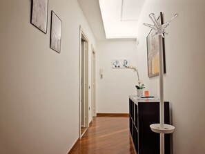Un pasillo con piso de madera, paredes blancas y una lámpara en el techo. Se observan cuadros en las paredes, un jarrón blanco con una orquídea y un perchero blanco.
