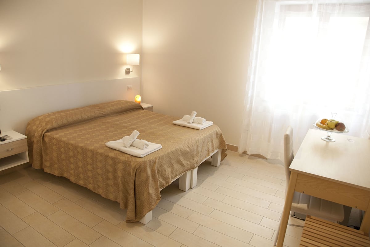 A hotel room with a bed, bedside table, and a small table with fruit and a glass.