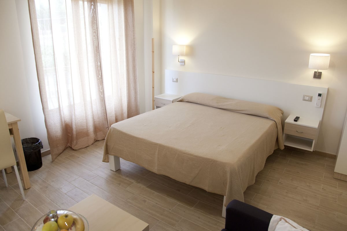 A hotel room with a bed, bedside tables, a nightstand, a lamp, a trash bin, and a table with a fruit bowl.