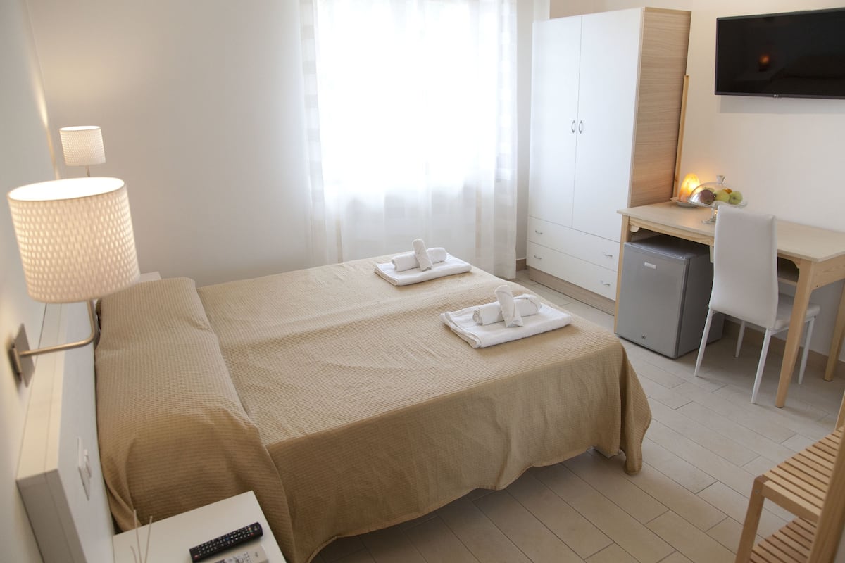 A hotel room with a bed, a desk, a chair, a lamp, and a TV.