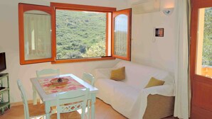 Ein Zimmer mit einer weißen Couch, einem kleinen Tisch mit floralem Tischläufer und zwei Stühlen. Zwei Fenster mit Holzrahmen, eines davon bietet Blick auf eine grüne Landschaft.