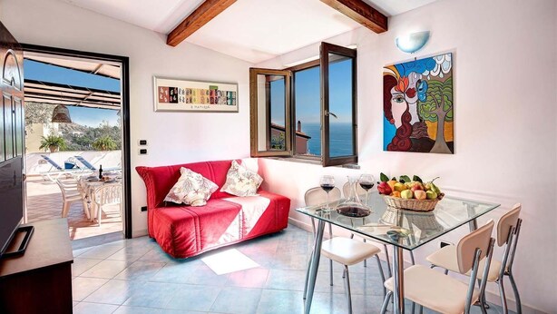 A modern living room with a red sofa, a glass dining table with chairs, and a view of the sea through large windows. - The Muse (Massa Lubrense)