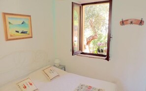 A bedroom with a bed, a window with a view of trees and flowers, a framed picture on the wall, and a small table with a lamp.