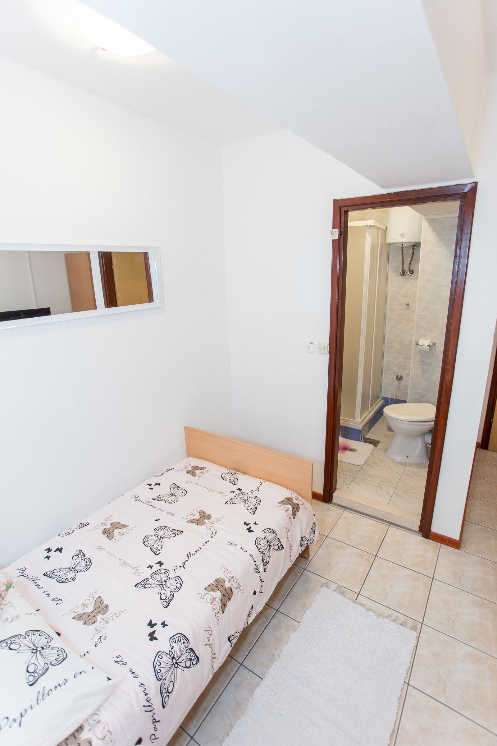 Habitación pequeña y limpia con una cama individual, un espejo y un baño visible a través de una puerta abierta.