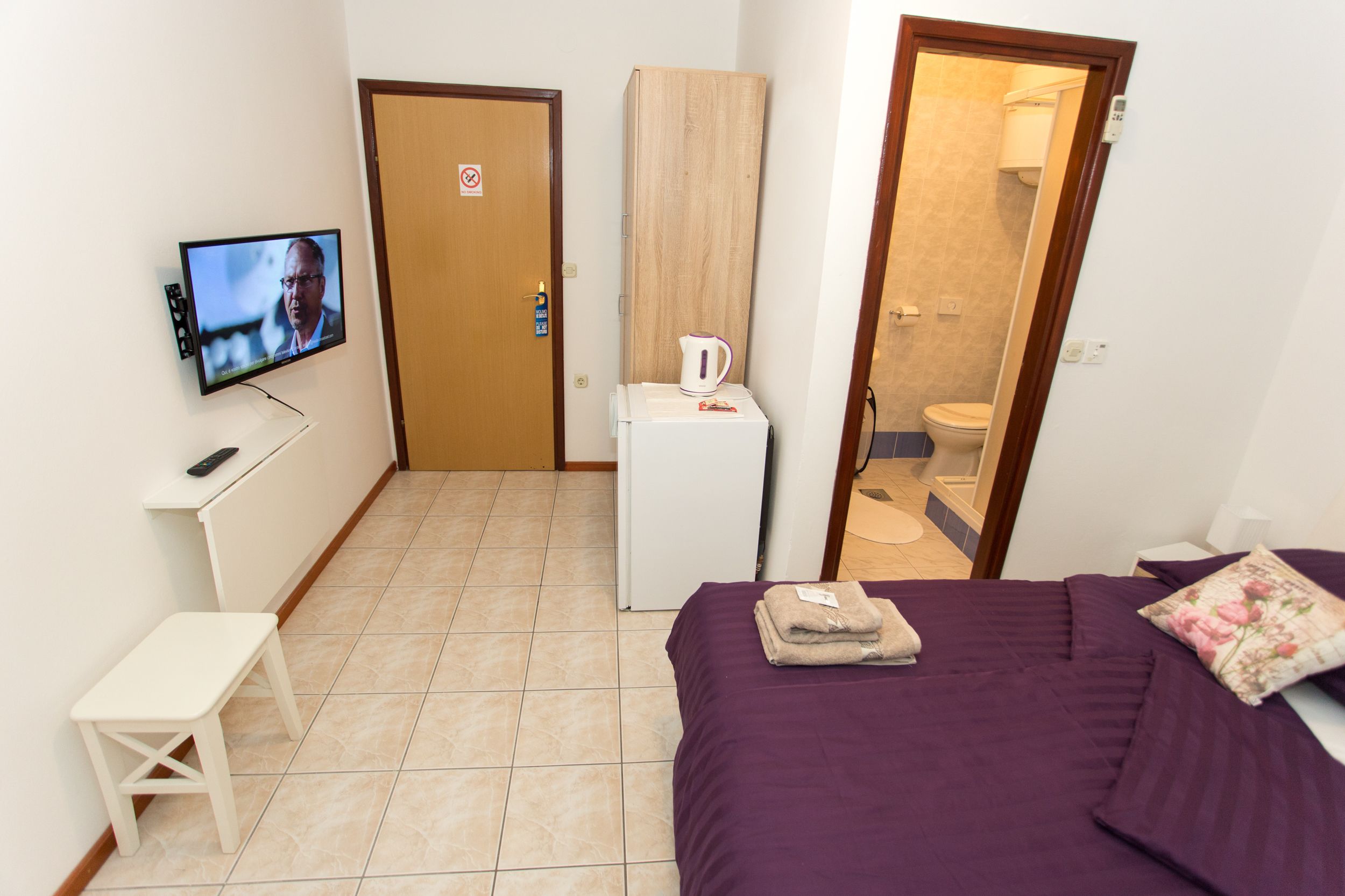 Een kleine, schone kamer met een bed, een televisie, een klein tafeltje en een deur naar een badkamer.