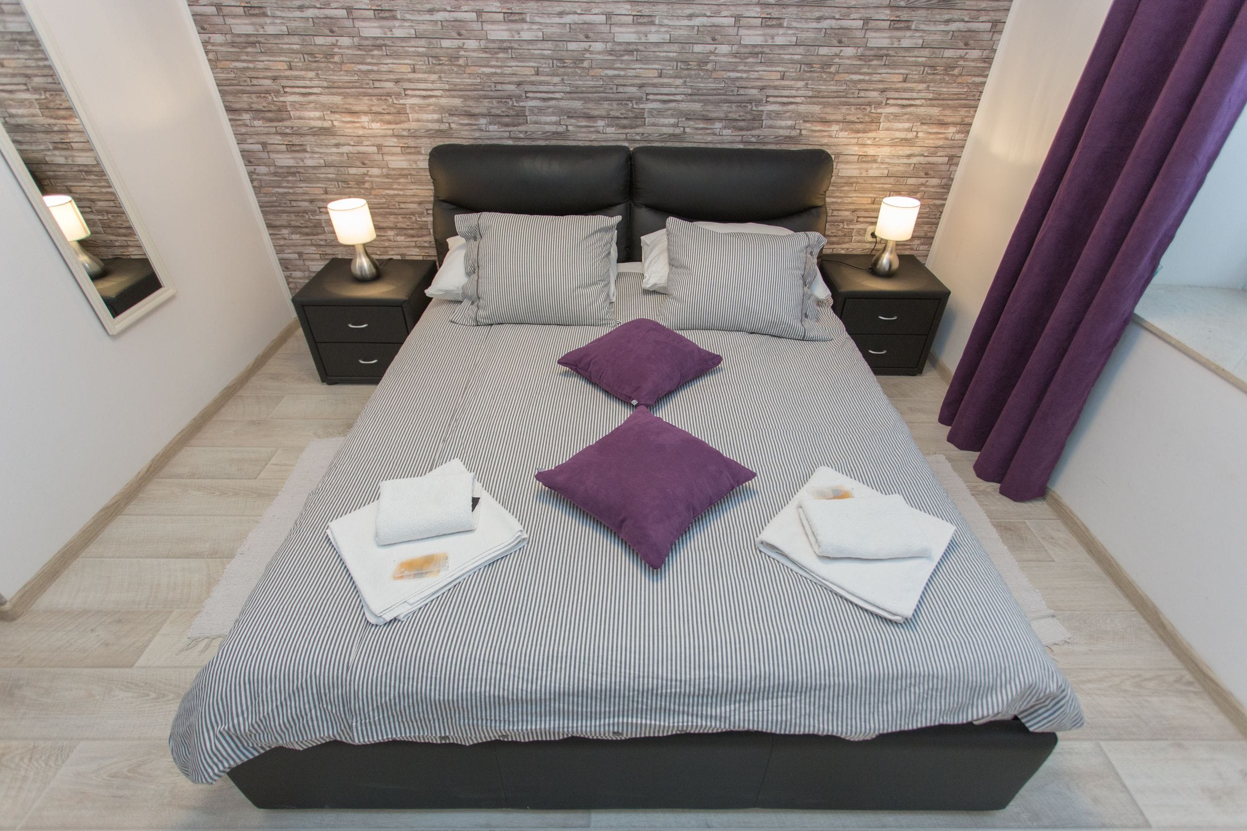 Une chambre moderne avec une tête de lit à motifs de briques, un grand lit avec une literie à rayures et deux coussins violets, deux tables de chevet avec des lampes et un rideau violet.