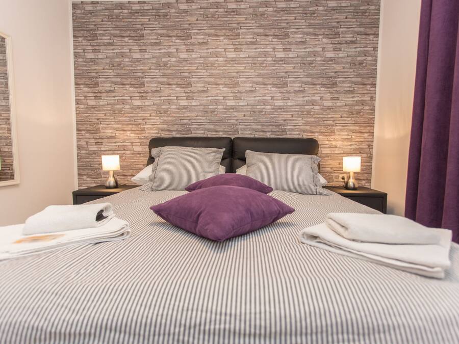 Habitació d'hotel amb paret de relleu de maó, un llit amb roba de llit a ratlles, dues taules de nit amb làmpades i almoines morades.