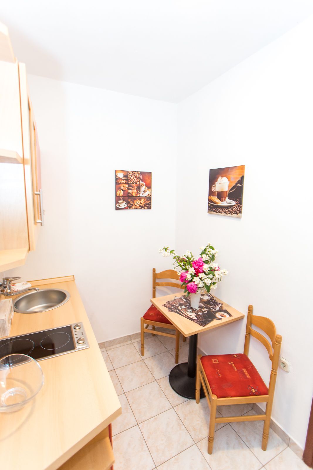 A small kitchenette with a stove, sink, and a dining table with chairs. There are two framed pictures on the wall.