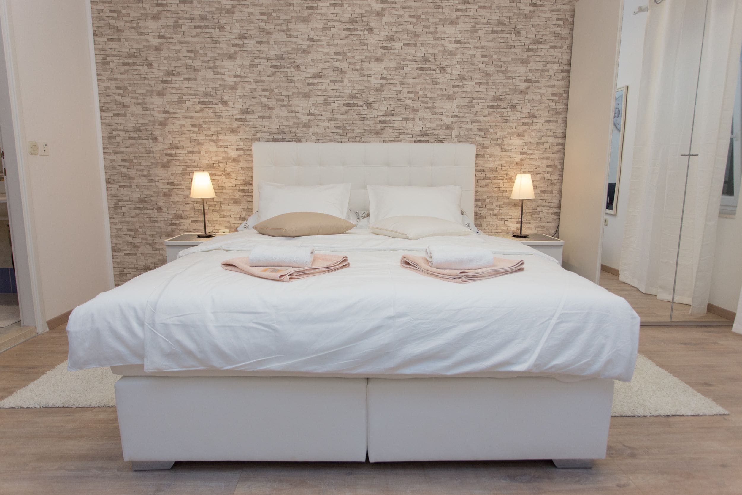 A modern bedroom with a brick-patterned wall, a white bed with two pillows, two bedside lamps, and a white rug.