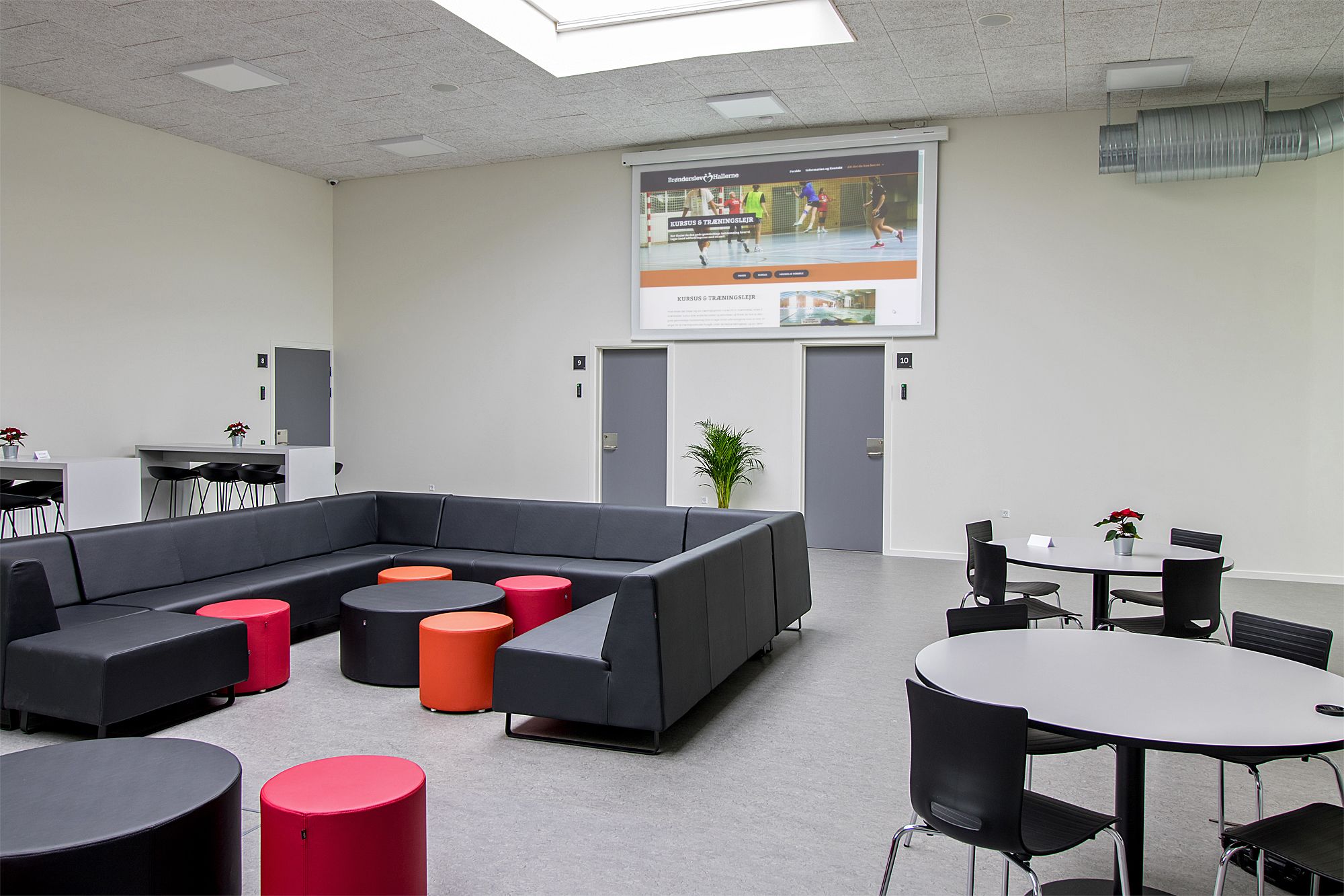 A modern conference room with a large screen, a sectional sofa, round tables, and chairs.