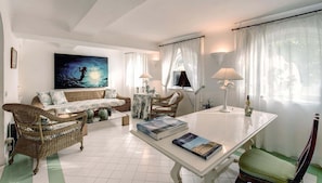 A spacious living room with a white sofa, wicker chairs, a white coffee table, and a dining table with books. - Green Coral (Capri)
