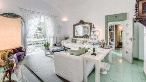 A spacious living room with a white sofa, a coffee table, and a large mirror. There is a bookshelf filled with books and a green door leading to another room. - Green Coral (Capri)