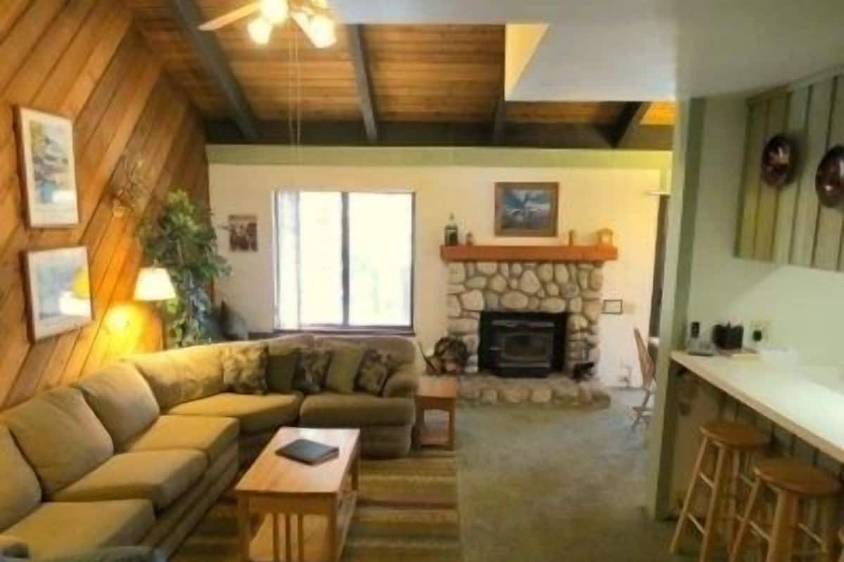 A cozy living room with a stone fireplace, wooden ceiling beams, and a large window.
