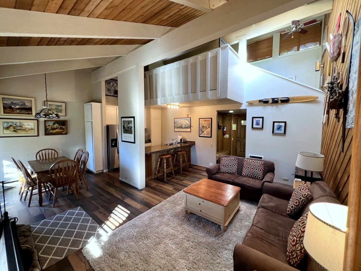 A spacious living area with a dining table, chairs, a sofa, and a coffee table. There is a kitchen area with a counter and stools. The room has wooden flooring and a high ceiling with exposed beams.