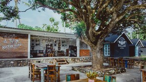 An outdoor seating area with rustic wooden tables and chairs, surrounded by a stone wall and a large tree. - Batan Sabo Cottage (Penida Island)