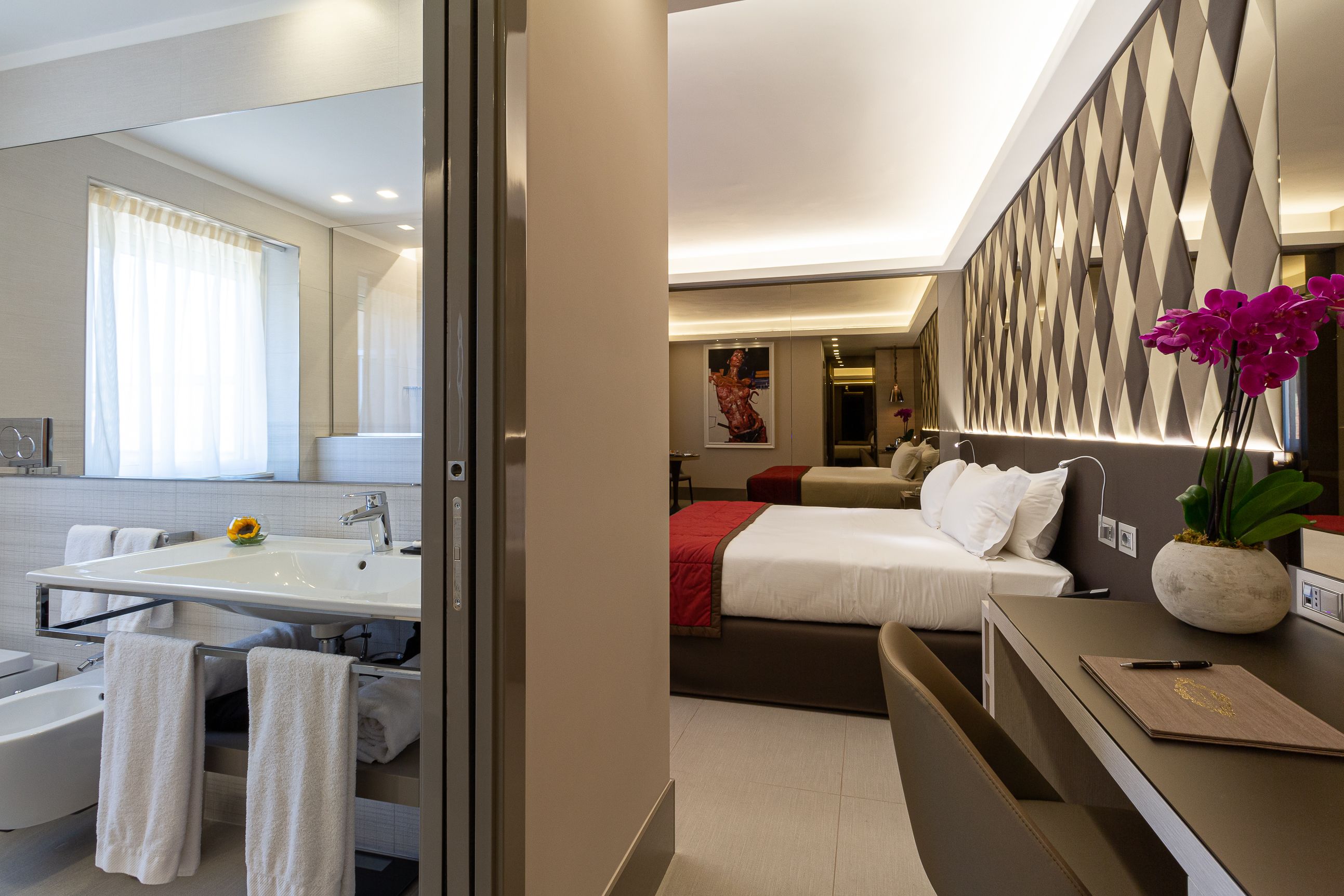 A modern hotel room with a large mirror, a bathroom with a sink and bathtub, a bed with a red blanket, and a desk with a vase of flowers.