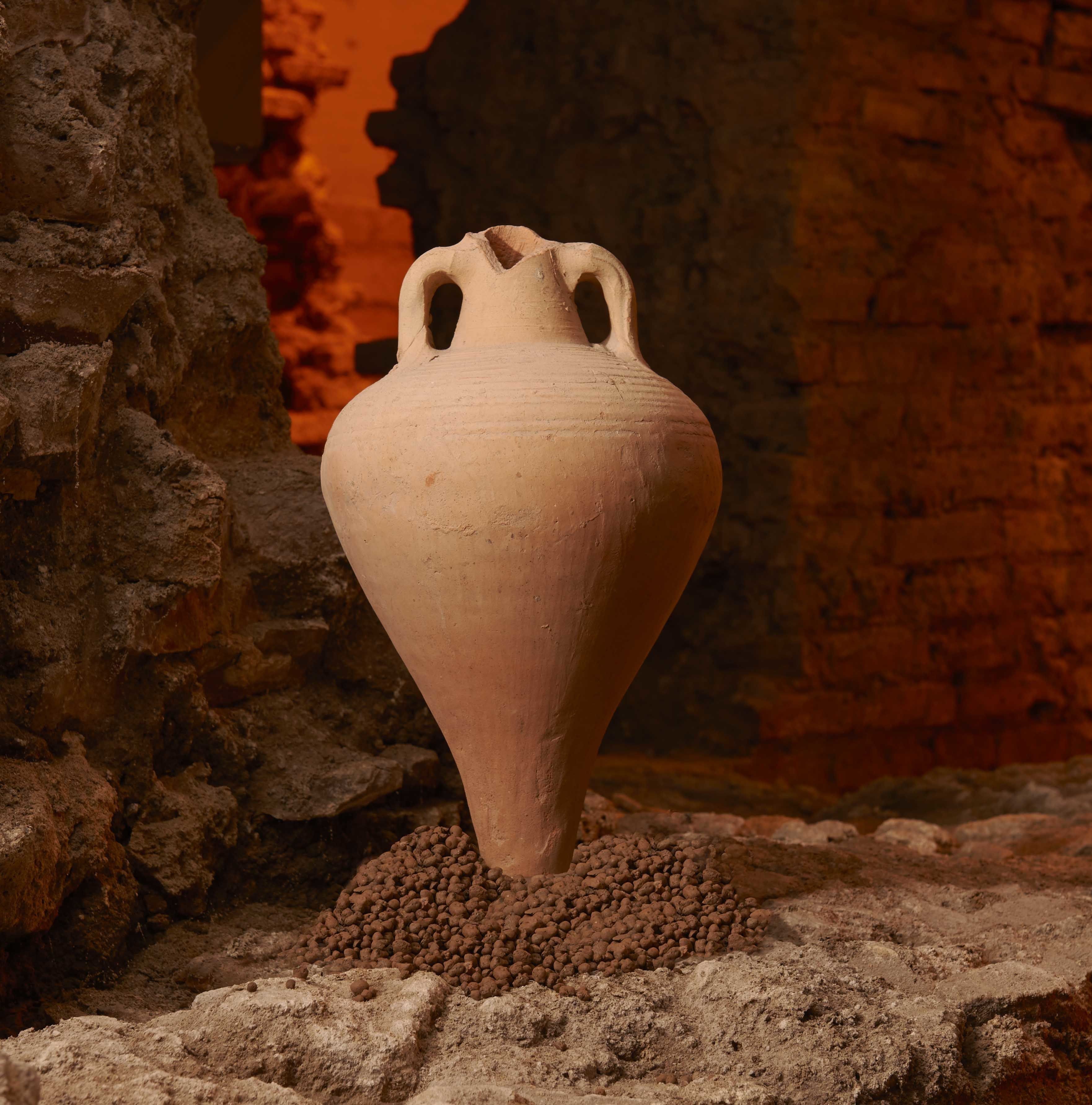 A terracotta vase with a narrow neck and two handles, placed on a sandy surface with a brick wall in the background.
