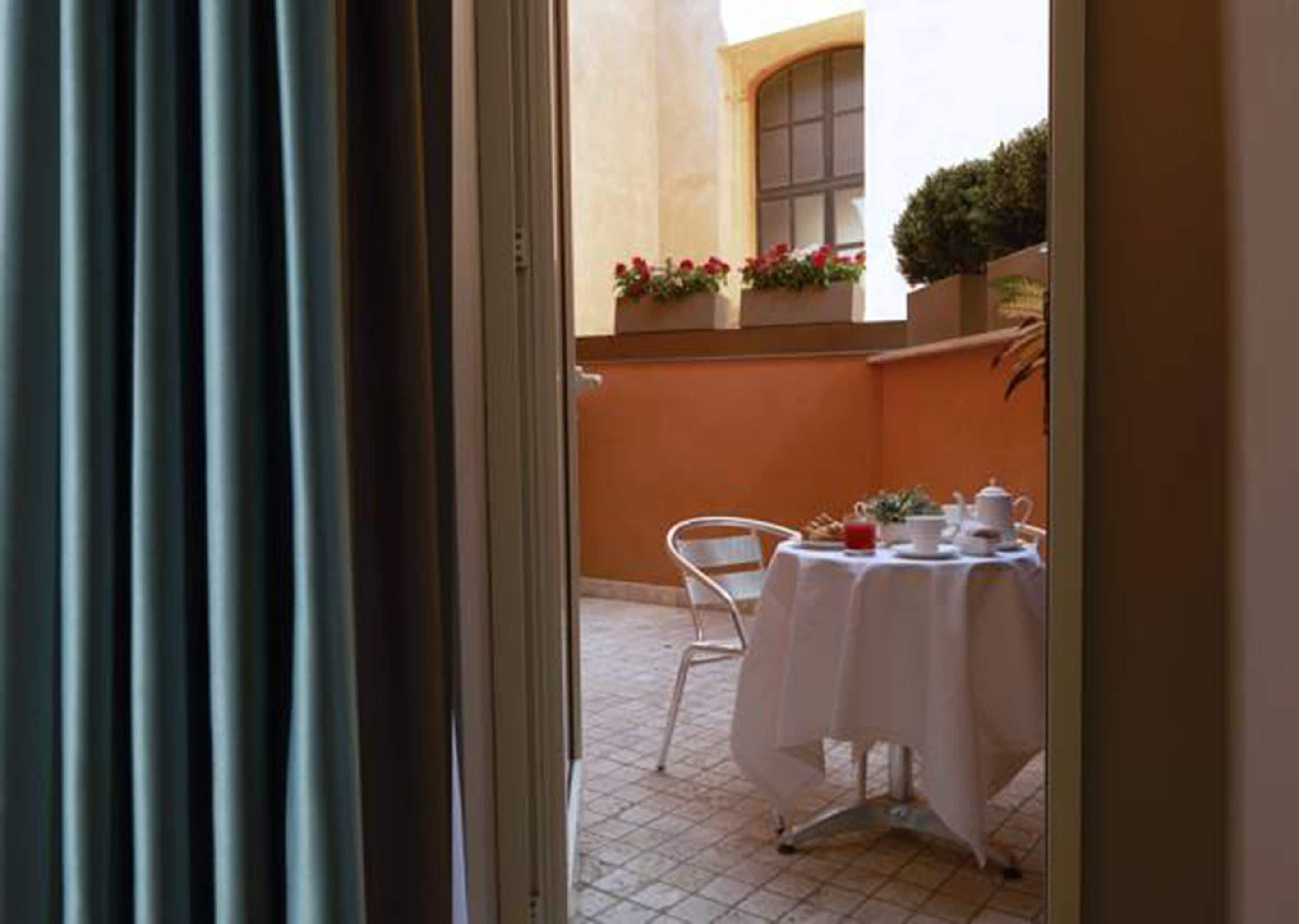 A room with a white table set for breakfast, a window with flowers, and a tiled floor.