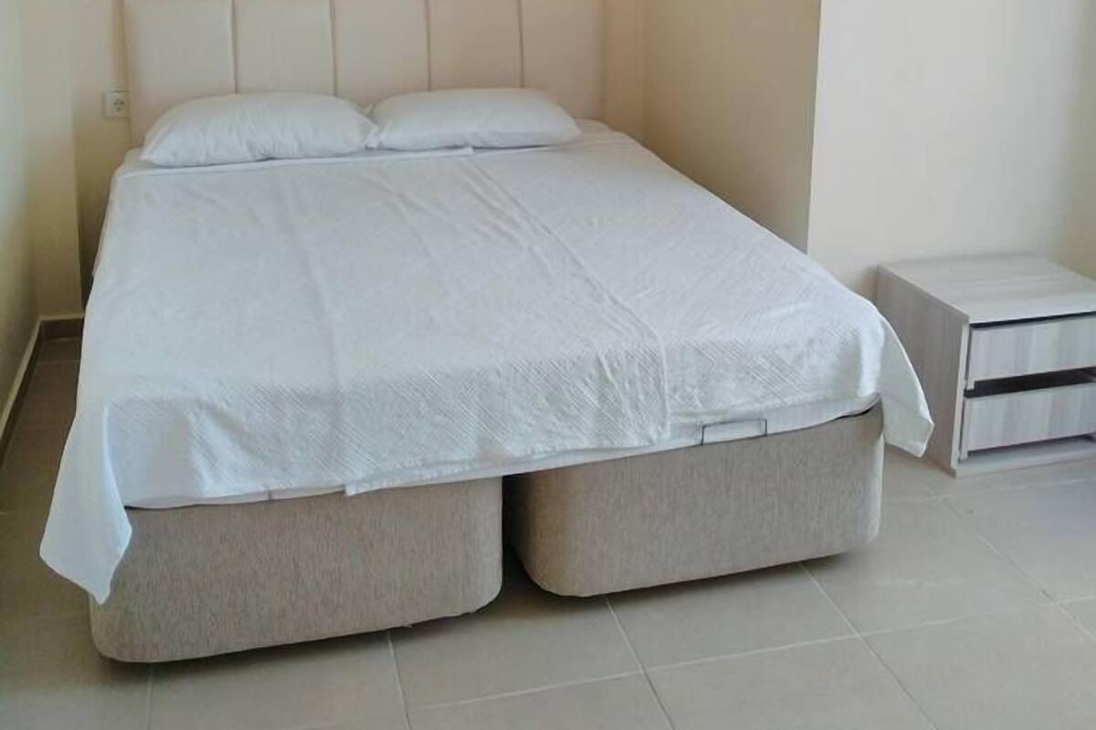 A bedroom with a bed, an air conditioning unit, a nightstand, and a lamp.