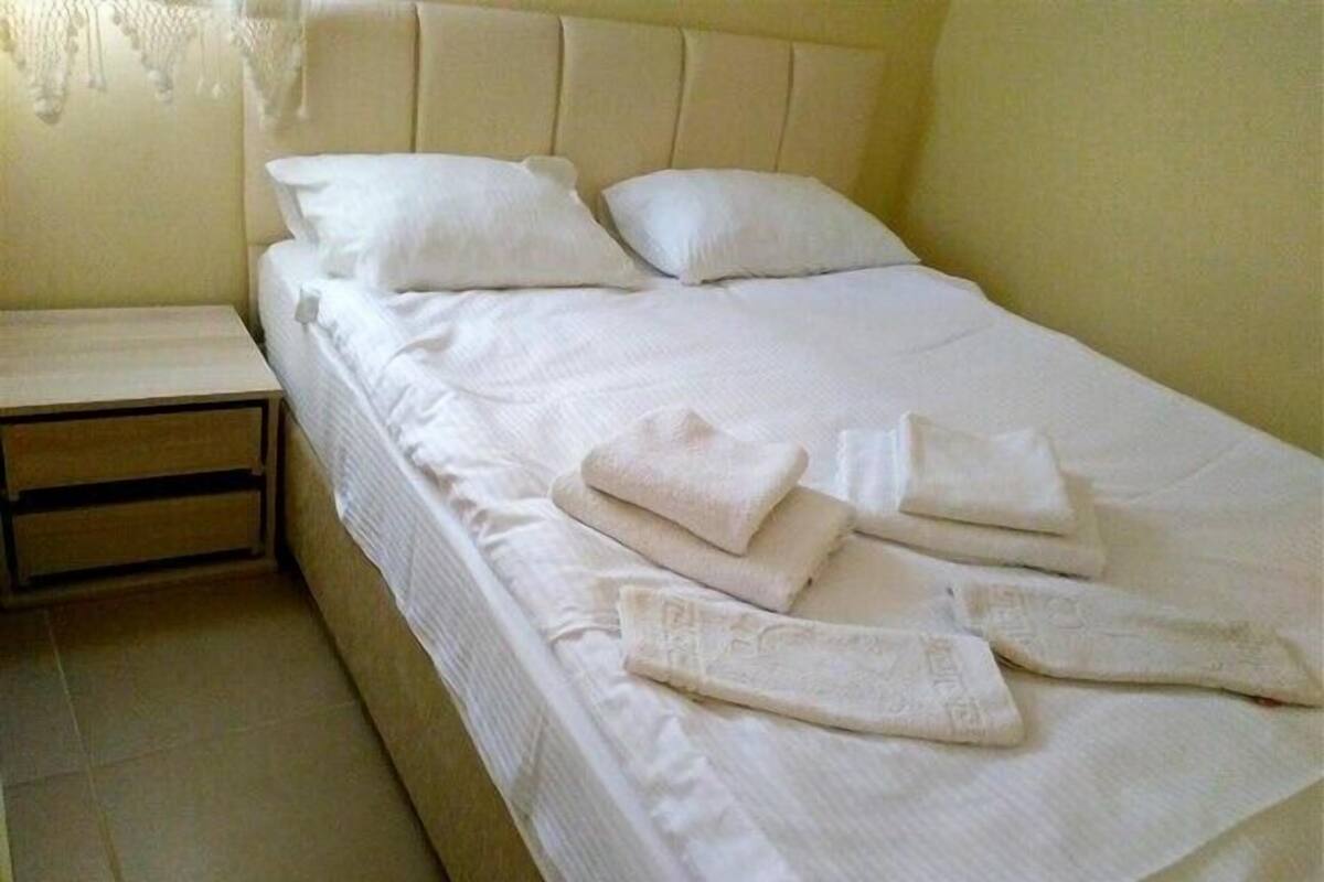 A neatly made bed with white linens and towels, a wooden nightstand, and a plain wall with a hanging light fixture.