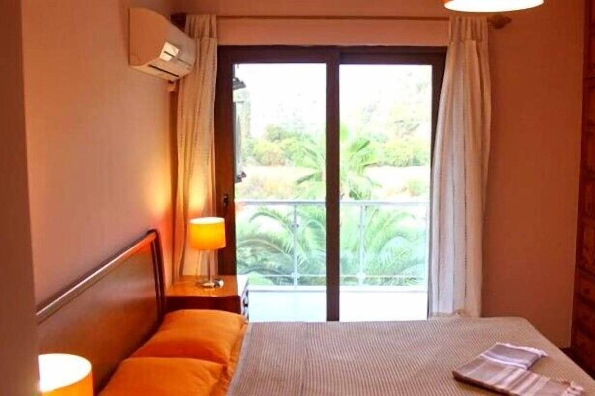 A hotel room with a bed, a nightstand with a lamp, an air conditioning unit, and a view of greenery through the window.