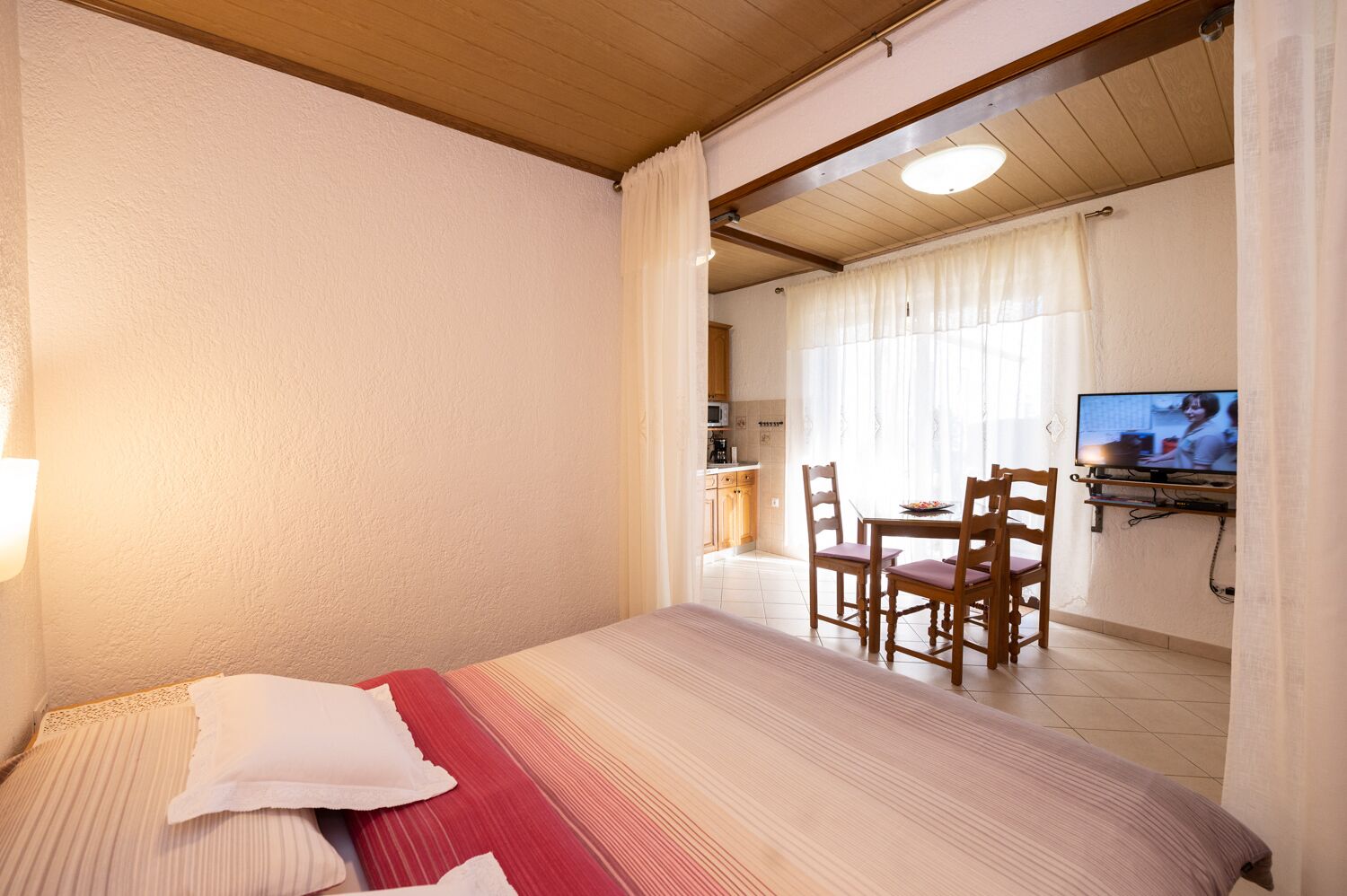 A hotel room with a bed, a dining area with chairs, a television, and a kitchen in the background.