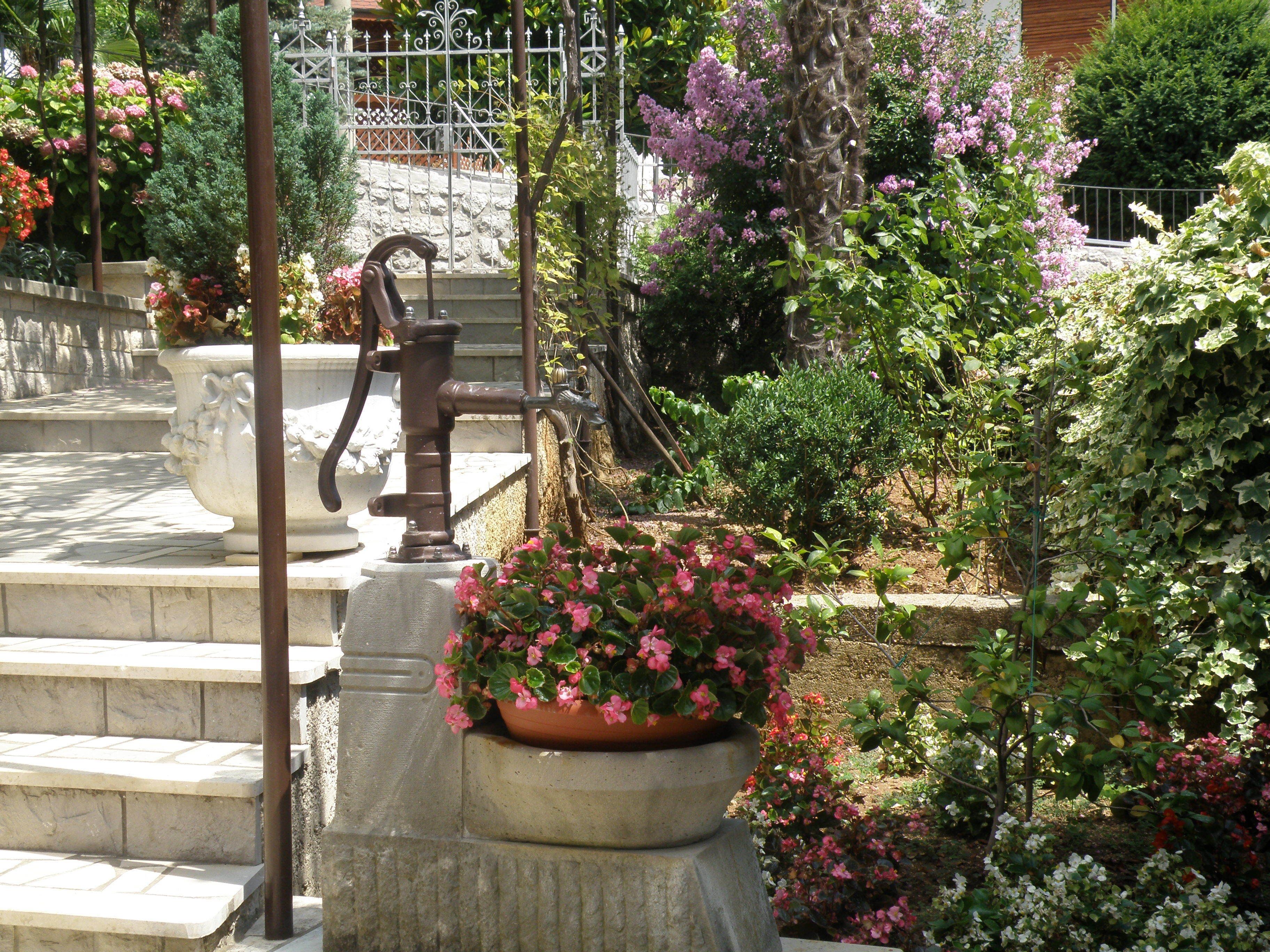 Um jardim com escadaria de pedra, cerca de metal e diversas plantas floridas.