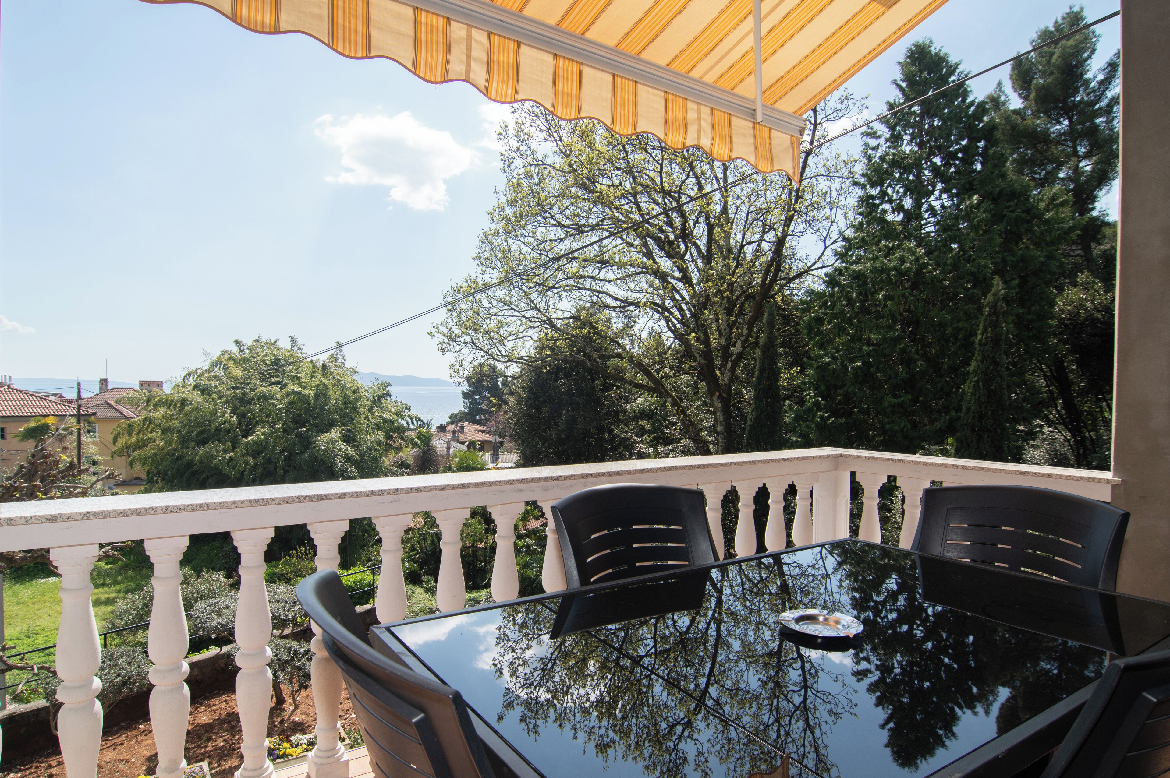 A balcony with a glass table and chairs, overlooking a garden and distant buildings.