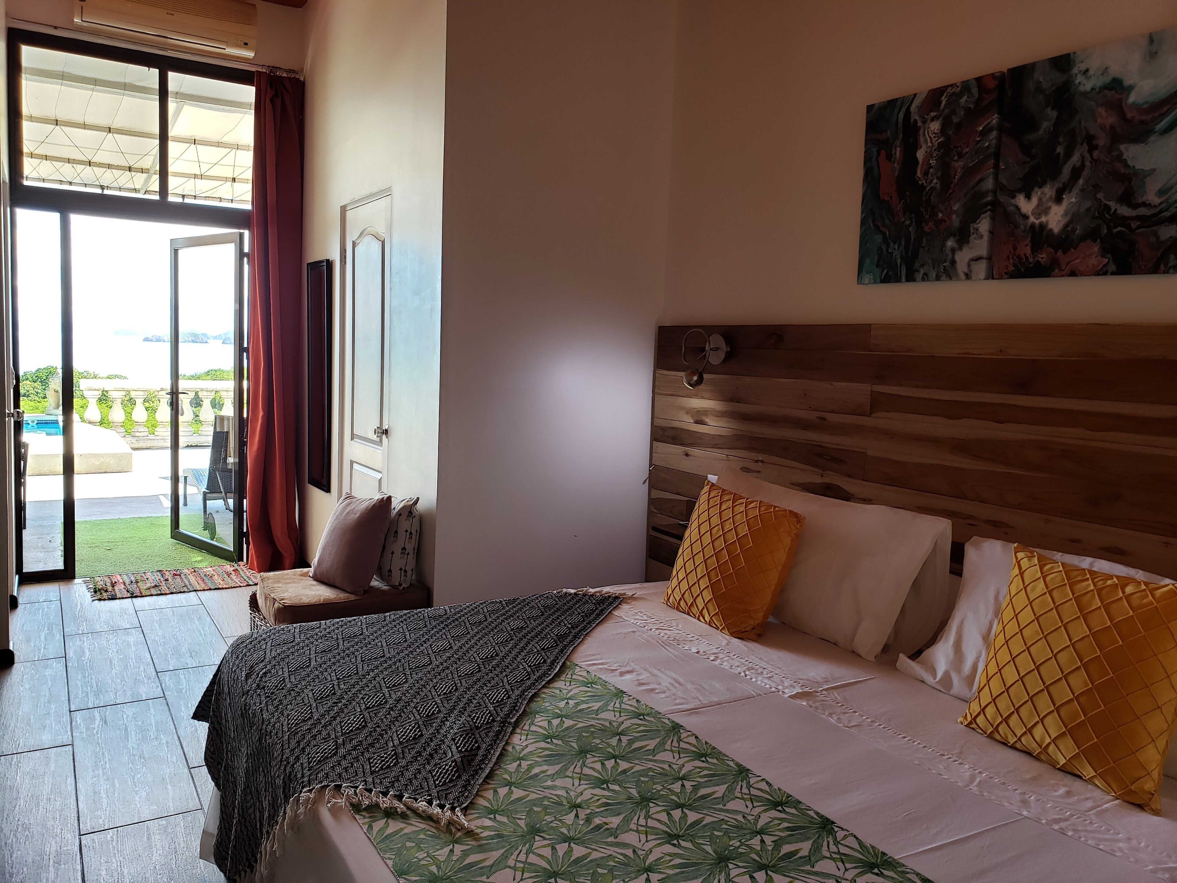 A bedroom with a bed, a wooden headboard, a painting, and a view of a pool and greenery through an open door.
