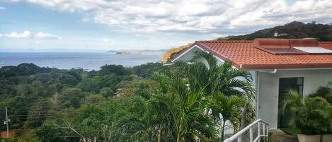 A coastal view with a building featuring a red-tiled roof, surrounded by lush greenery and a clear sky.