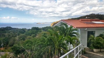 A coastal view with a building featuring a red-tiled roof, surrounded by lush greenery and a clear sky.