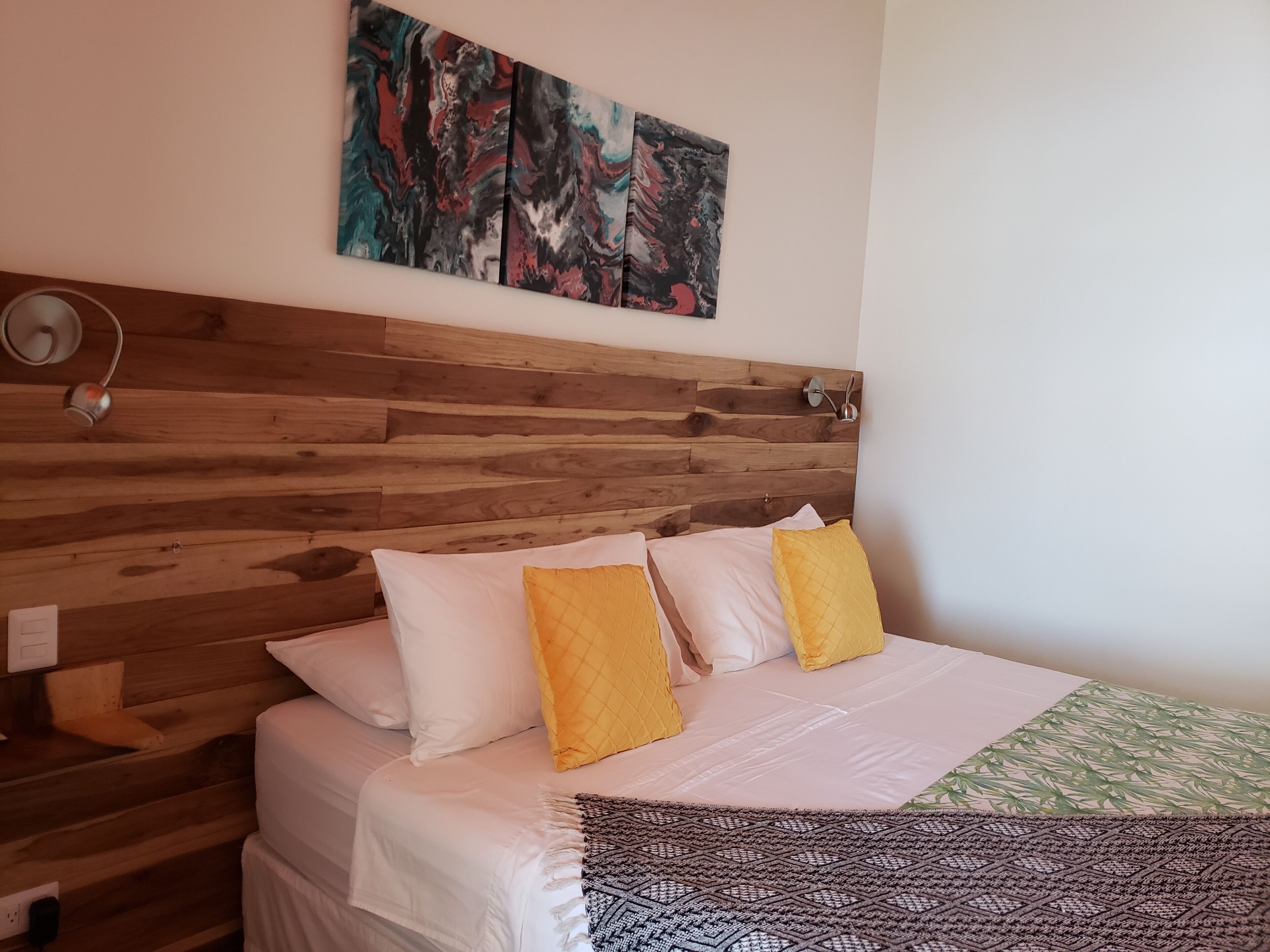 Une chambre à coucher avec une tête de lit en bois, du linge de lit blanc et un tableau abstrait coloré au-dessus du lit.